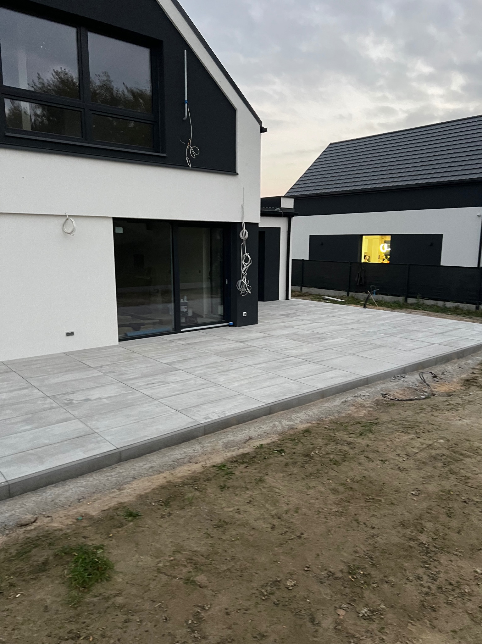 Nowoczesny taras z szarych płyt betonowych, obrzeża z kostki, widok na dom z czarno-białą elewacją i okablowaniem zewnętrznym, trawnik w tle.