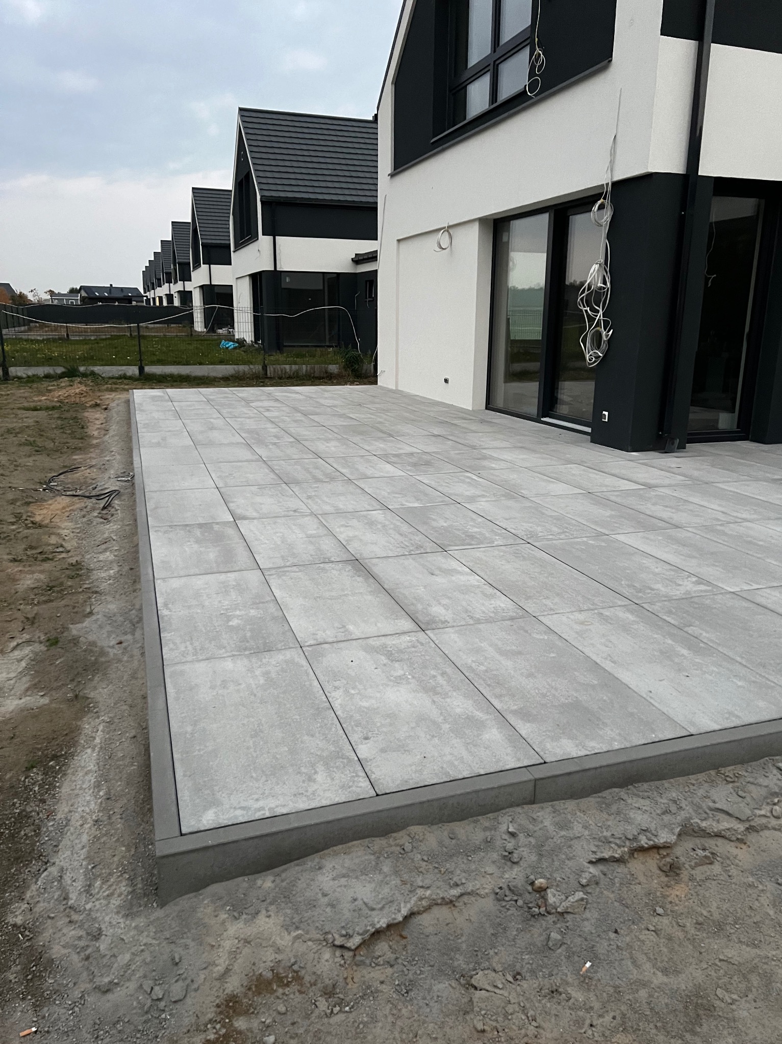 Ułożona, szara kostka brukowa na posesji przed szeregiem nowoczesnych domów z czarno-białą elewacją, widoczne kable zwisające przy ścianie budynku.