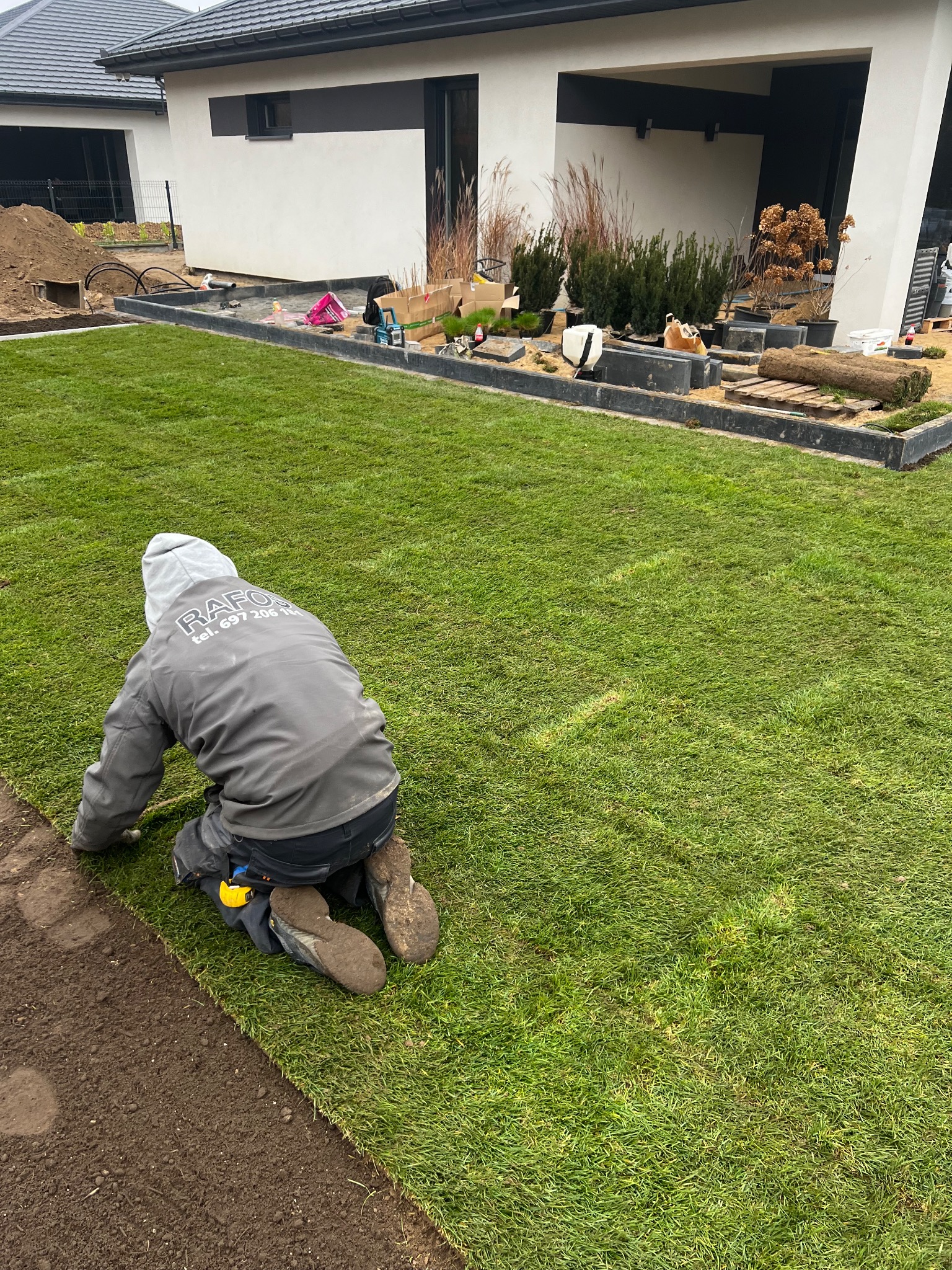 Pracownik w szarej bluzie z kapturem i logo firmy układa rolki trawy na przygotowanym podłożu, w tle nowoczesny dom z elementami dekoracyjnymi i roślinnością.