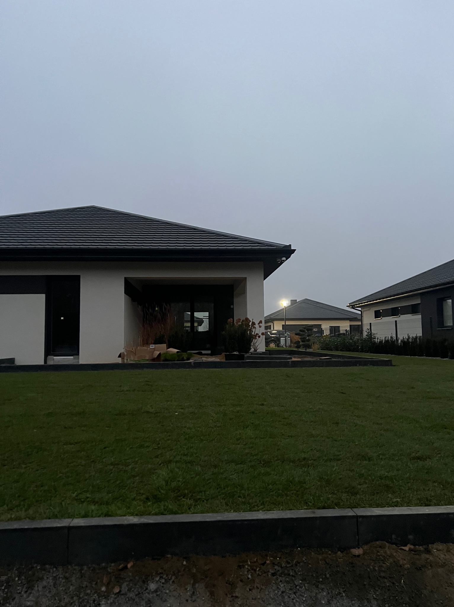 Nowoczesny dom jednorodzinny z zadbanym trawnikiem i elementami dekoracyjnymi z kostki brukowej, widok z poziomu gruntu w pochmurny dzień.