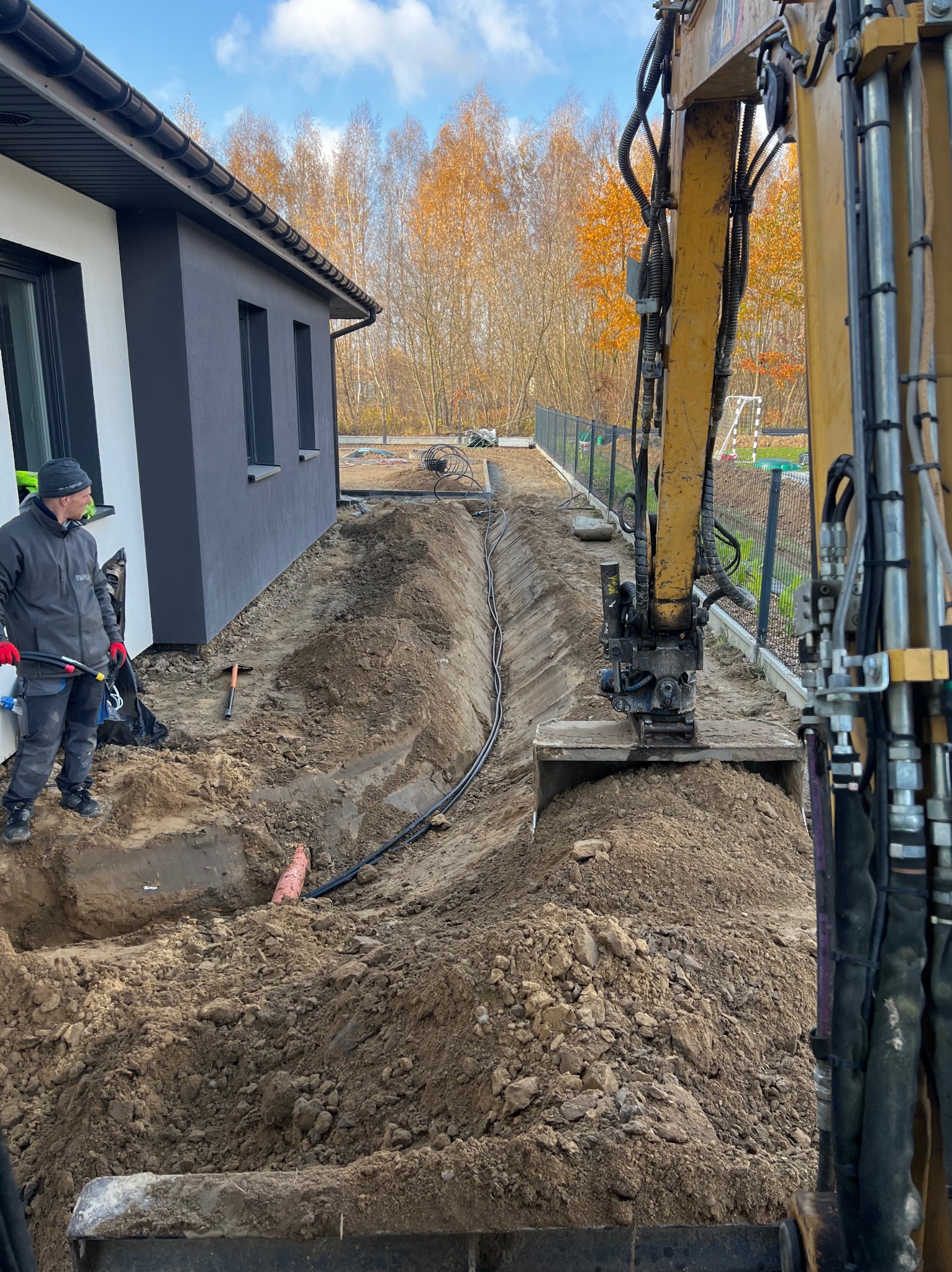 Żółta koparka w trakcie prac ziemnych, wykopany wąski rów wzdłuż budynku z ułożonymi kablami, pracownik w oddali przygląda się pracom.