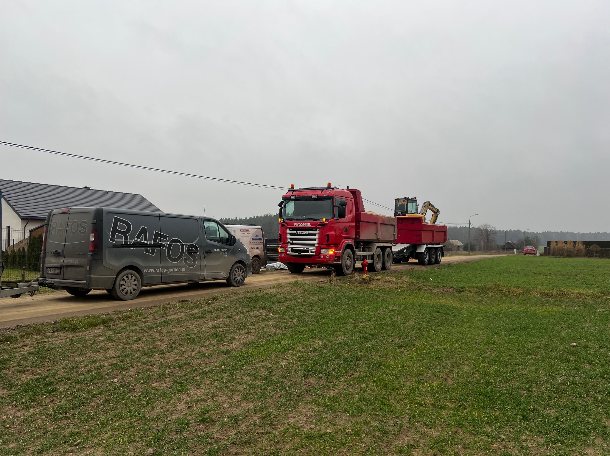 Czerwona ciężarówka Scania z przyczepą i koparką na platformie, zaparkowana obok szarego busa z logo firmy 'RAFOS' na tle zielonego pola i pochmurnego nieba.