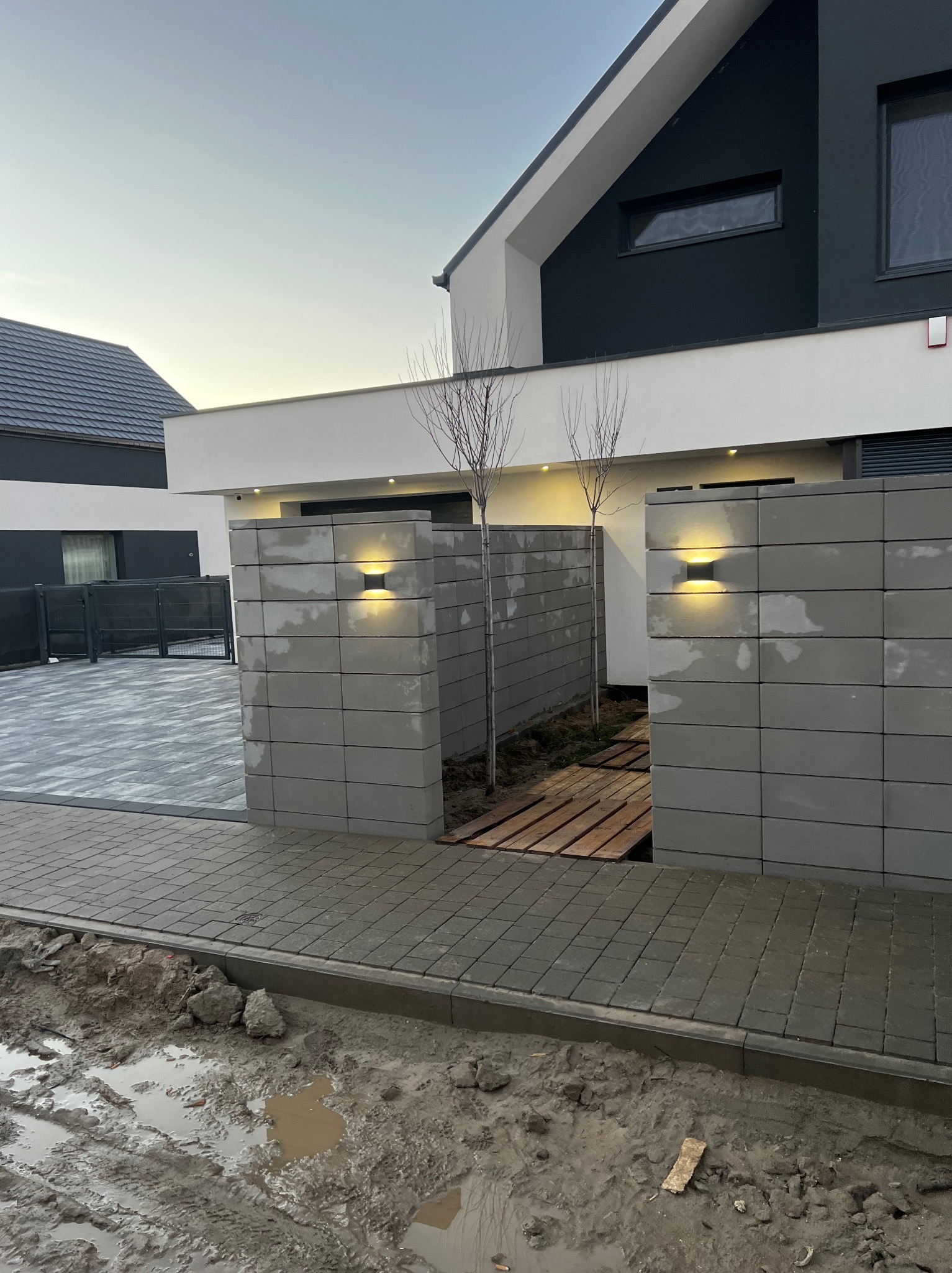 Nowoczesne ogrodzenie z betonowych bloczków z oświetleniem zewnętrznym, widok na posesję z kostką brukową i fragment błotnistego terenu przed posesją.