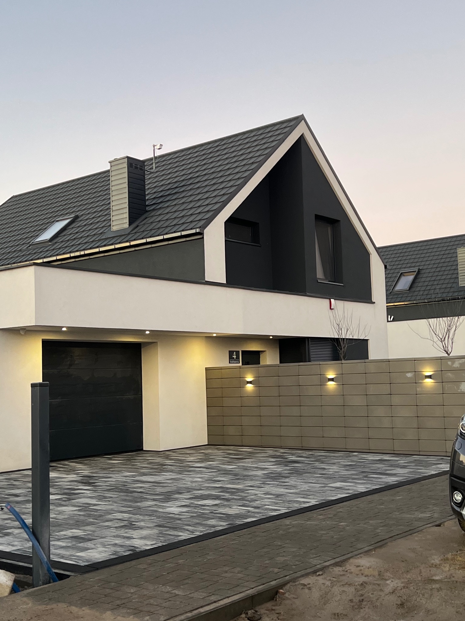 Nowoczesny dom jednorodzinny z garażem i podjazdem wyłożonym szaro-białą kostką brukową ułożoną we wzór szachownicy, oświetlony lampami zewnętrznymi.