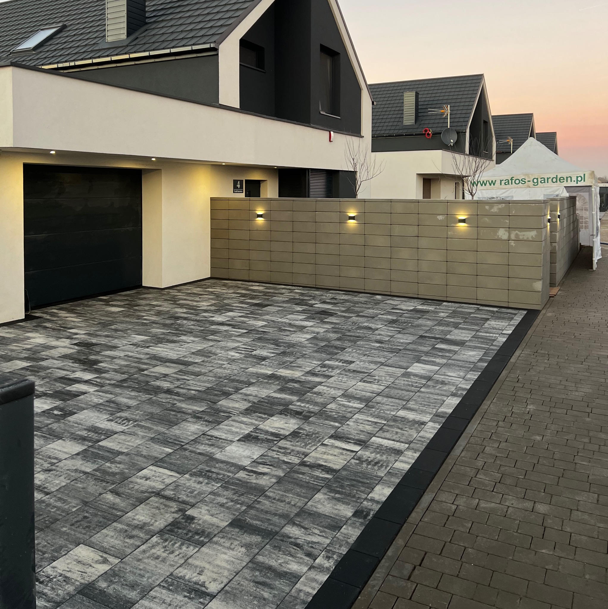 Ułożona kostka brukowa w odcieniach szarości na podjeździe nowoczesnego domu z czarną bramą garażową i oświetlonym murem, widoczny fragment namiotu z logo firmy.