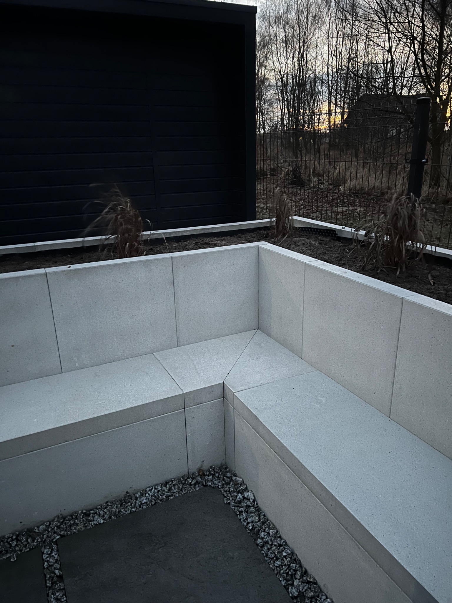 Nowoczesny narożny mur z jasnoszarej kostki, tworzący siedzisko w ogrodzie, otoczony drobnym żwirem i elementami roślinności.