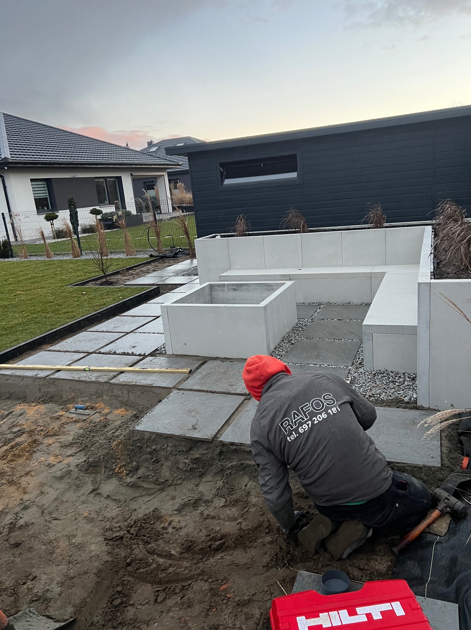 Pracownik firmy Rafos w czerwonej czapce układa szare płyty chodnikowe na podsypce piaskowej, widoczny poziomica, młotek i skrzynka narzędziowa Hilti, w tle nowoczesny dom z ogrodem.