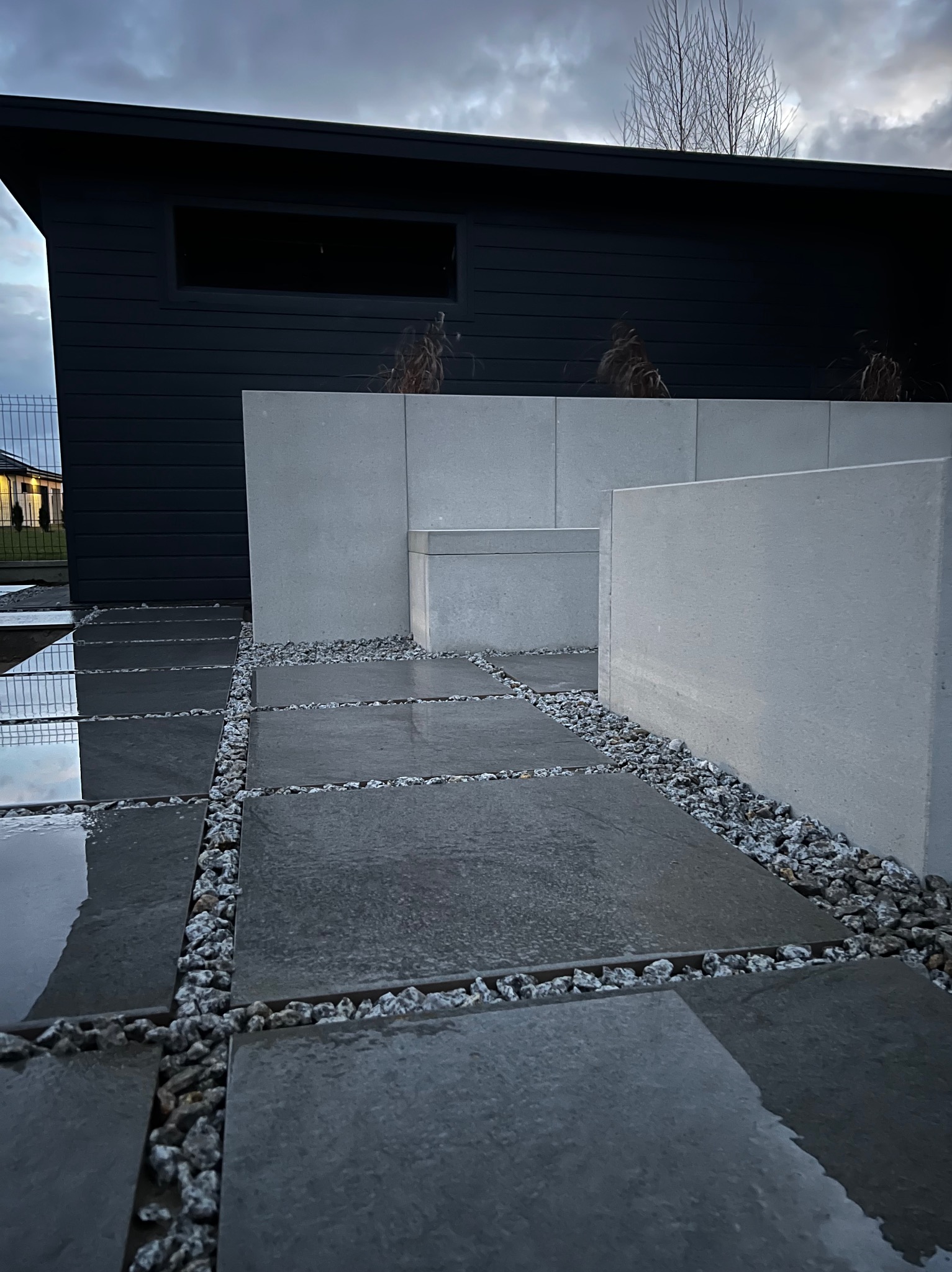 Nowoczesna nawierzchnia z ciemnych płyt betonowych oddzielonych białym grysem, przylegająca do minimalistycznej, ciemnej bryły budynku z jasnymi, betonowymi elementami dekoracyjnymi.