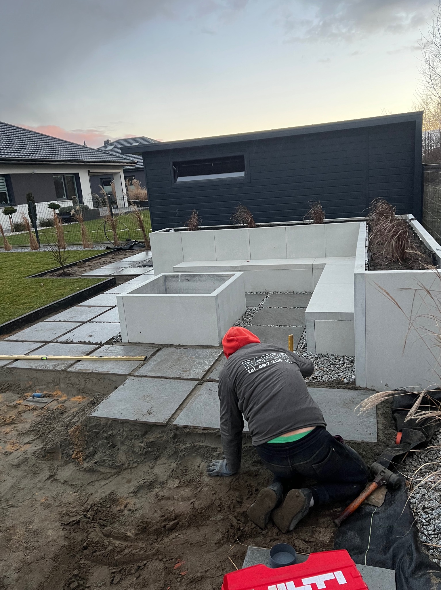 Układanie szarych płyt chodnikowych na podsypce piaskowej, widoczny poziomica, fragment ogrodu z trawami ozdobnymi i nowoczesną architekturą ogrodową w tle, pracownik w ubrudzonych butach...