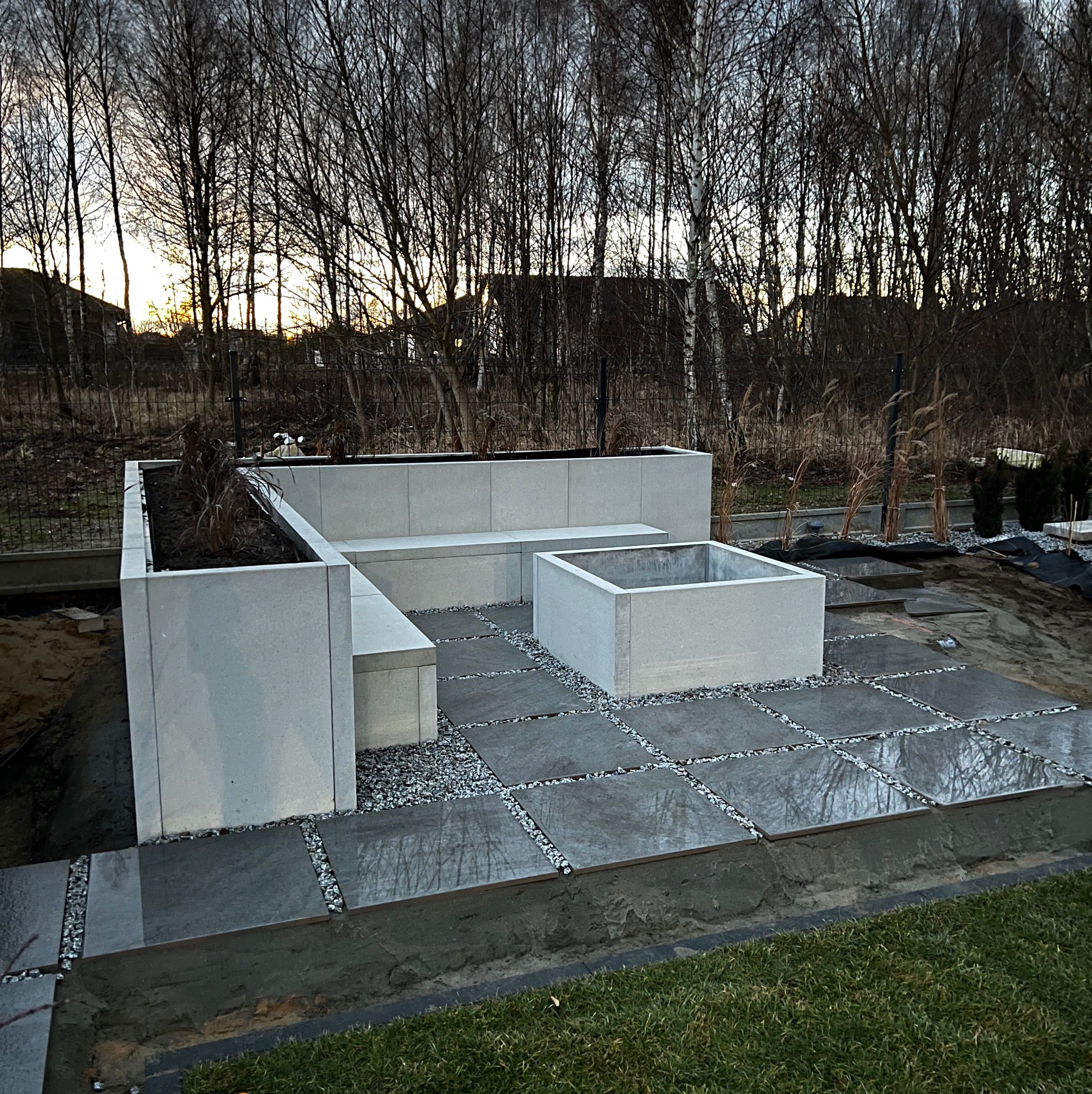 Nowoczesna aranżacja przestrzeni zewnętrznej z betonowych płyt i elementów siedziskowych, wypełniona drobnym żwirem, na tle drzew bez liści o zachodzie słońca.