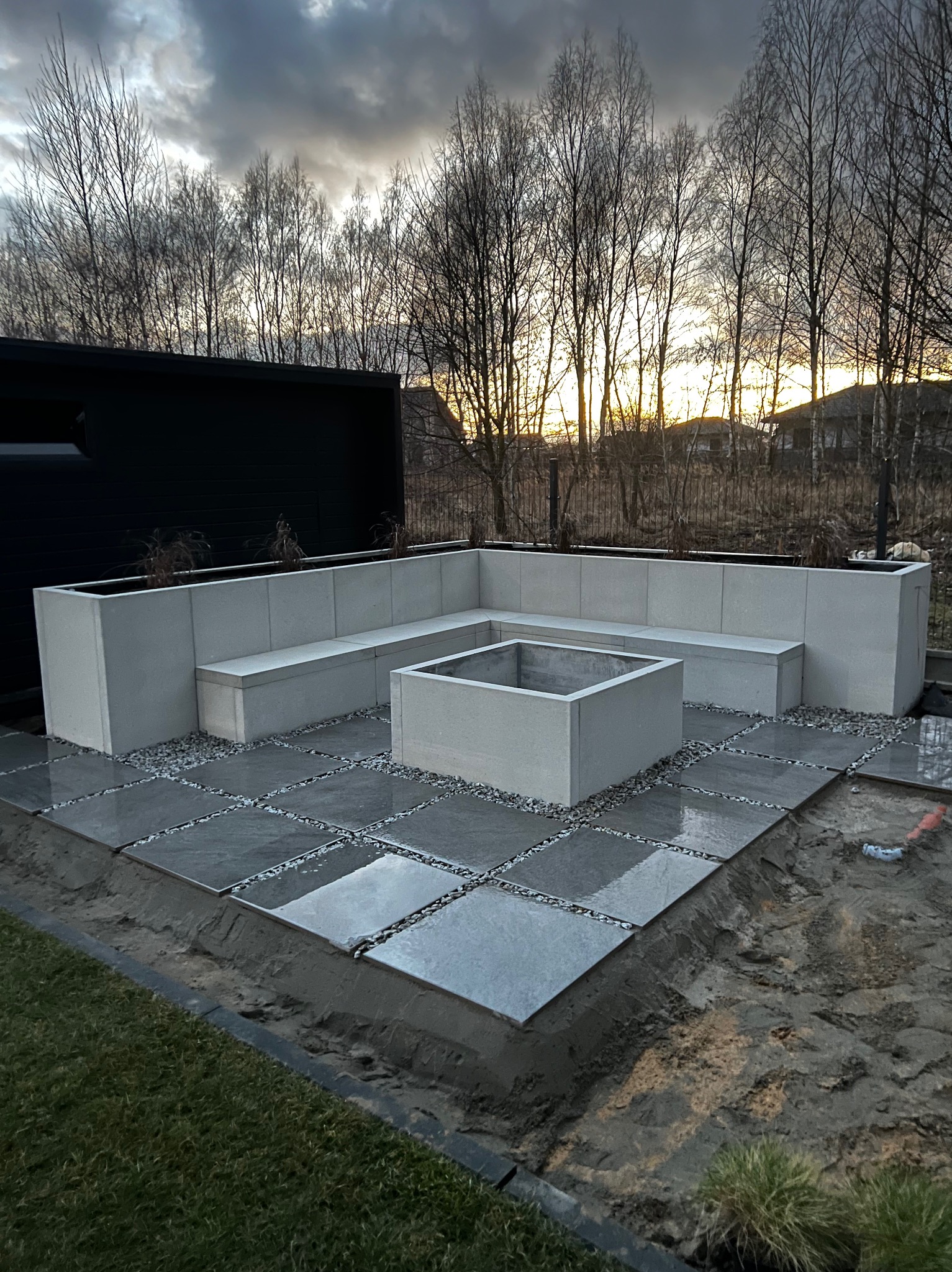 Nowoczesna aranżacja przestrzeni zewnętrznej z szarych płyt chodnikowych oddzielonych drobnym żwirem, otoczona betonową ławką w kształcie litery L i centralnym paleniskiem, na tle zachodzącego...