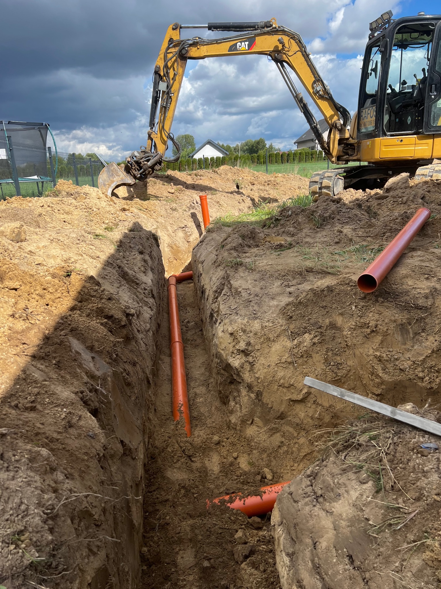Żółta koparka CAT przy wykopie z pomarańczowymi rurami kanalizacyjnymi. Widoczne narzędzia i dom w tle. Pochmurne niebo.
