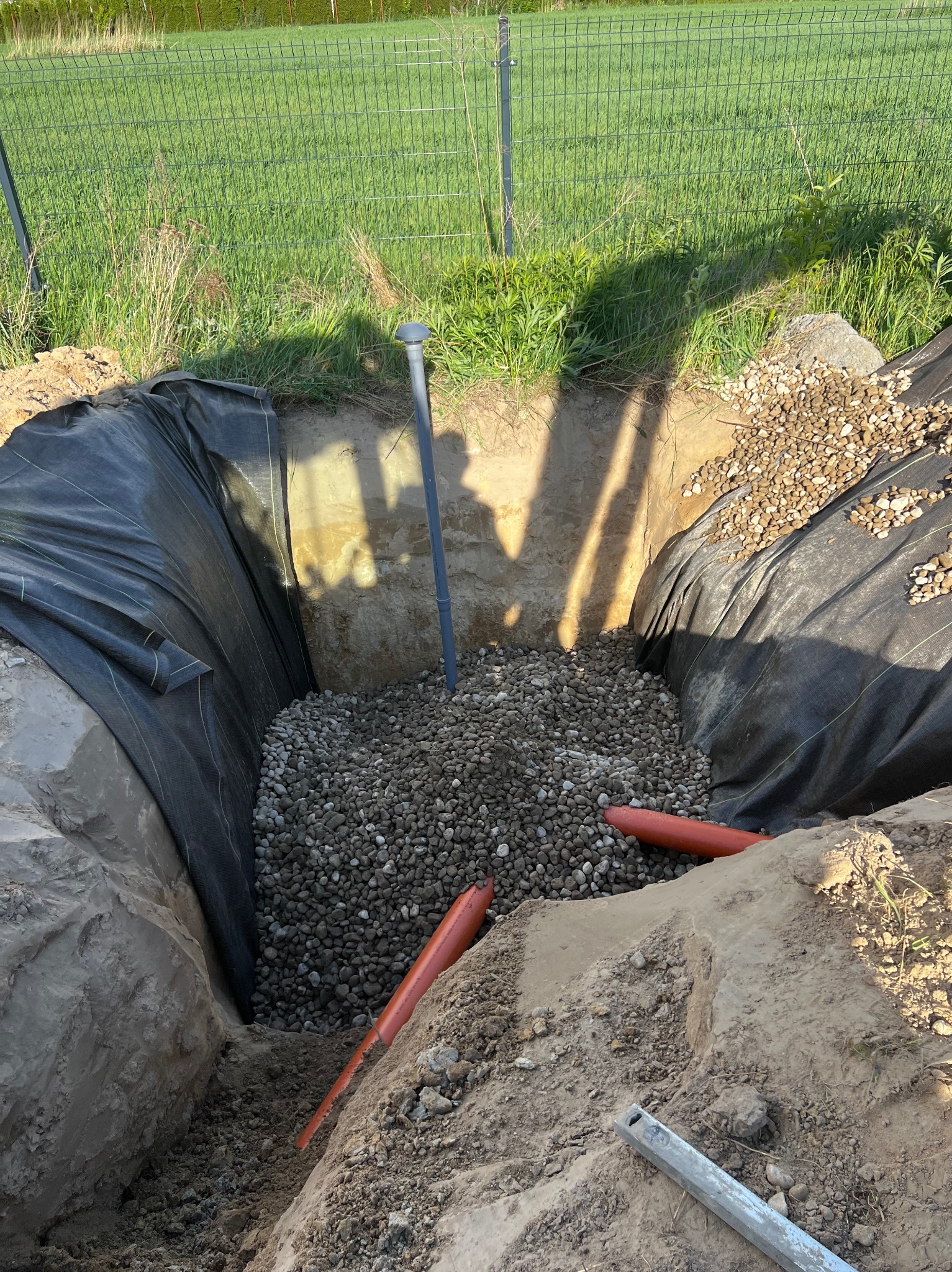 Wykop ziemny wyłożony czarną folią, wypełniony kamieniami, z wystającymi rurami drenażowymi i pionowym przewodem wentylacyjnym, na tle zielonego trawnika i ogrodzenia.
