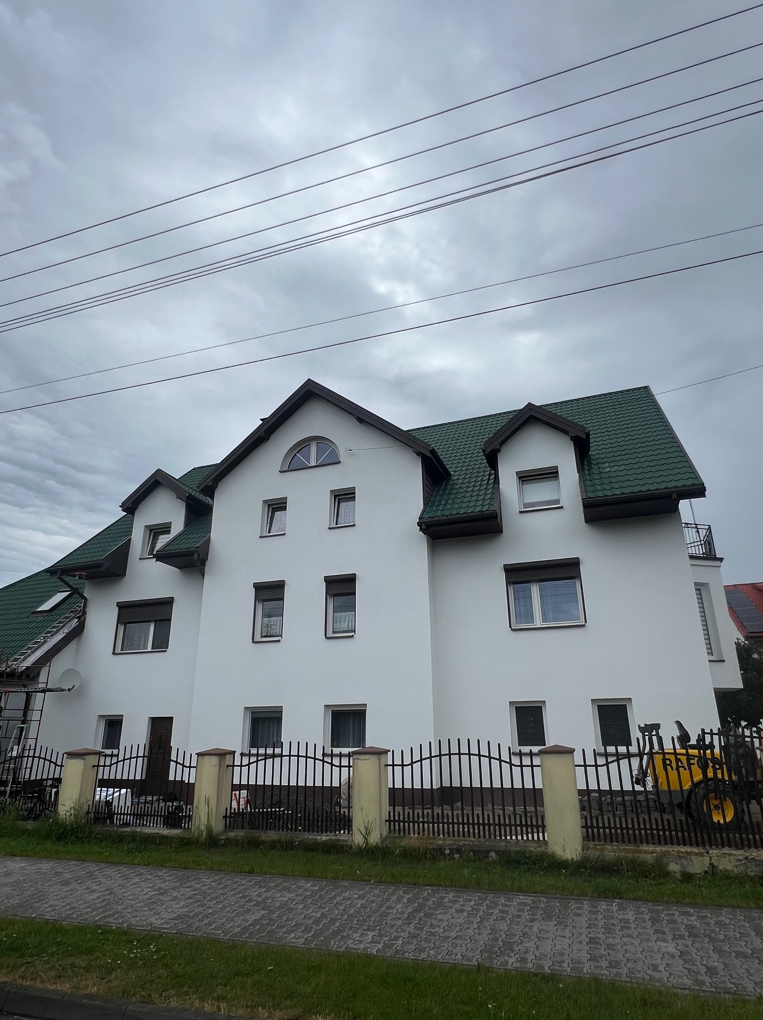 Biały, dwupiętrowy dom z zielonym dachem, widziany z poziomu chodnika. Metalowe ogrodzenie z betonowymi słupkami. Pochmurne niebo w tle. Widoczne przewody elektryczne.