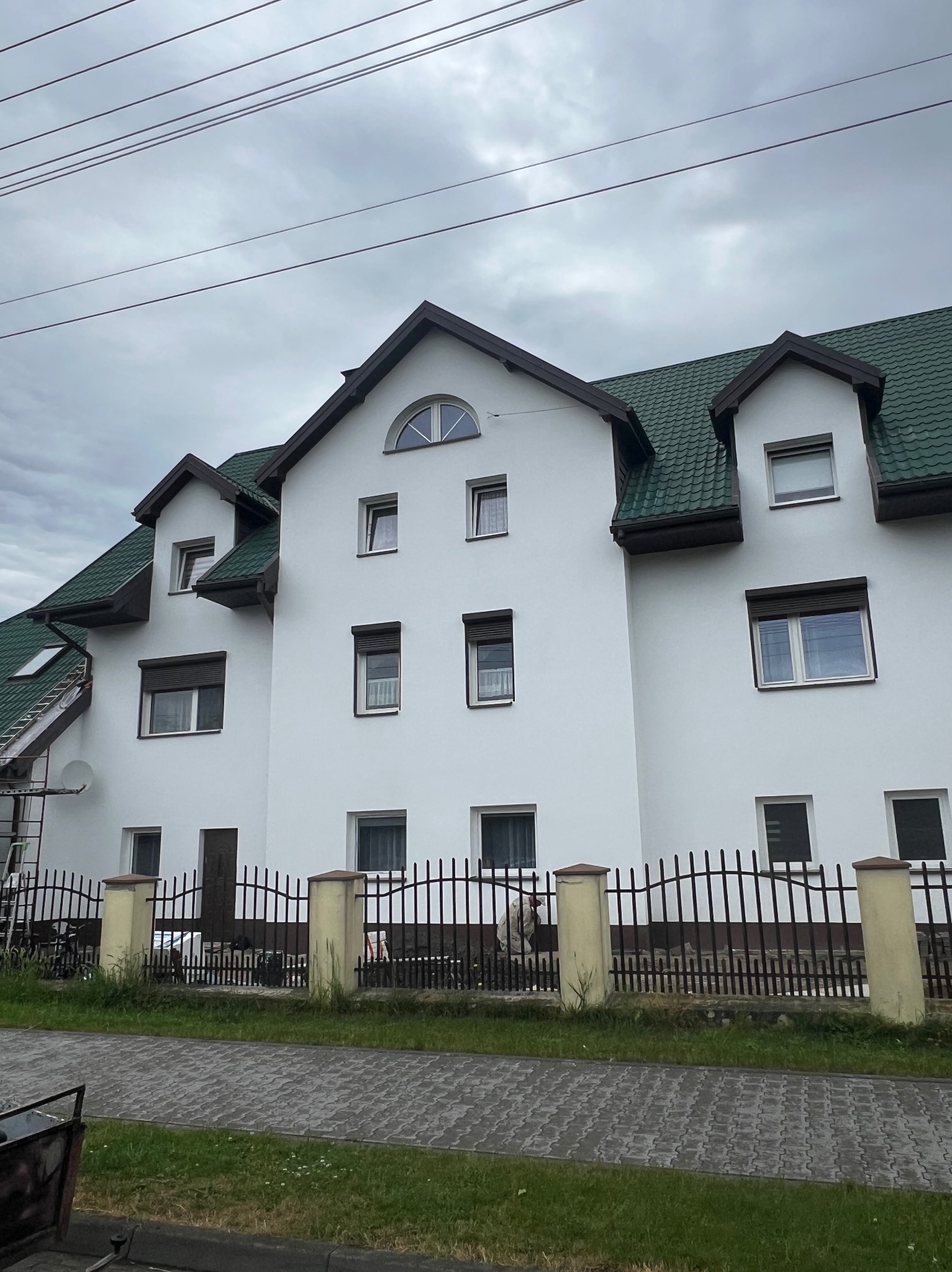 Biały budynek po renowacji w Sieradzu z zielonym dachem i ciemną stolarką okienną, ogrodzony kutym płotem. Widok z poziomu chodnika.