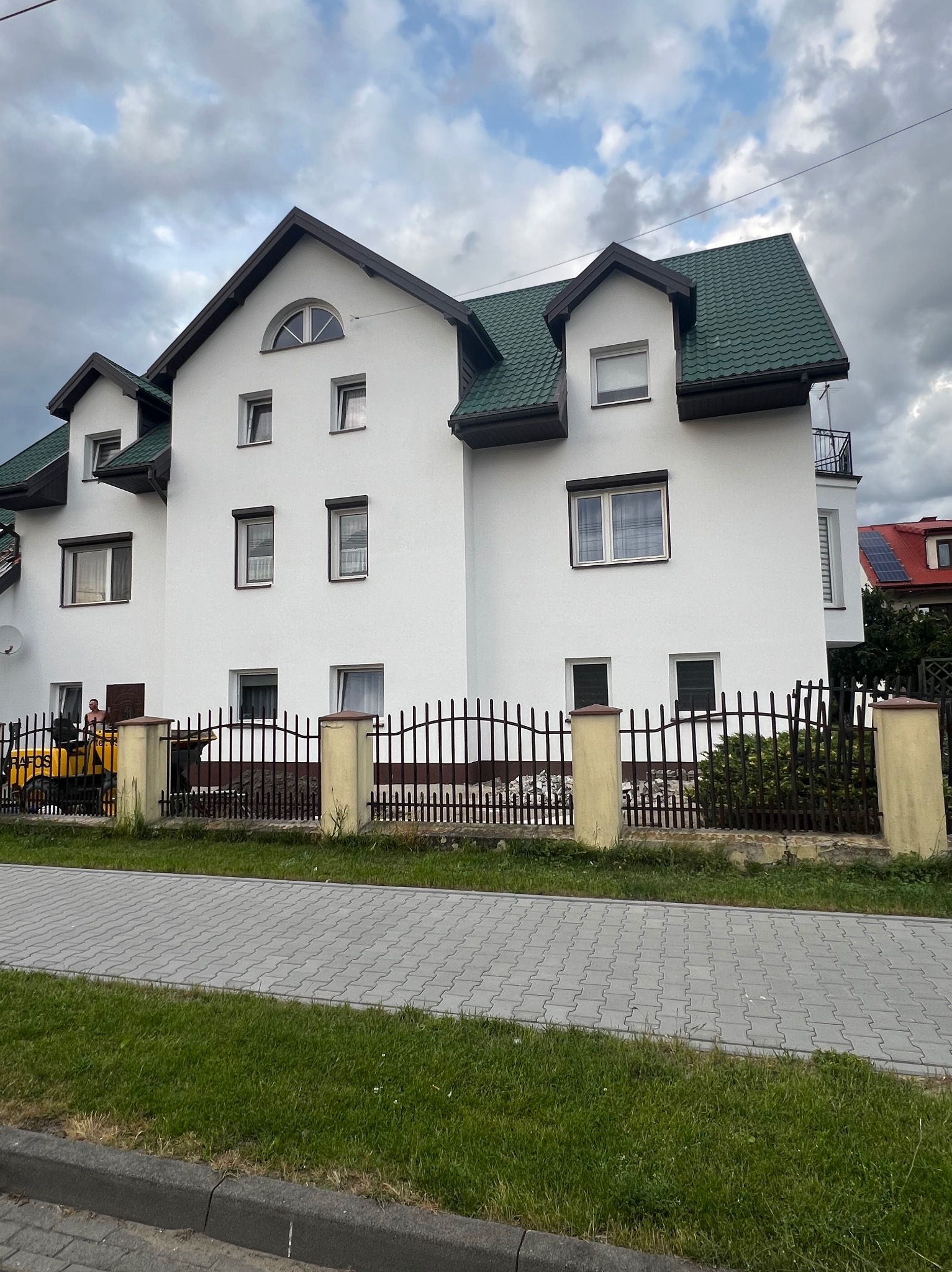Biały, trzypiętrowy dom z zielonym dachem w stylu dworkowym, ogrodzony czarnym płotem z żółtymi słupkami. Widoczny fragment chodnika z kostki brukowej i maszyna budowlana.