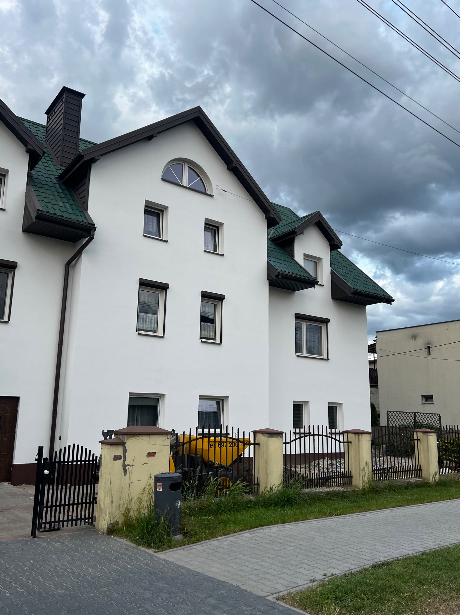 Biały dom z zielonym dachem, widoczny z poziomu chodnika. W tle pochmurne niebo i słupy elektryczne. Żółty sprzęt budowlany za ogrodzeniem.