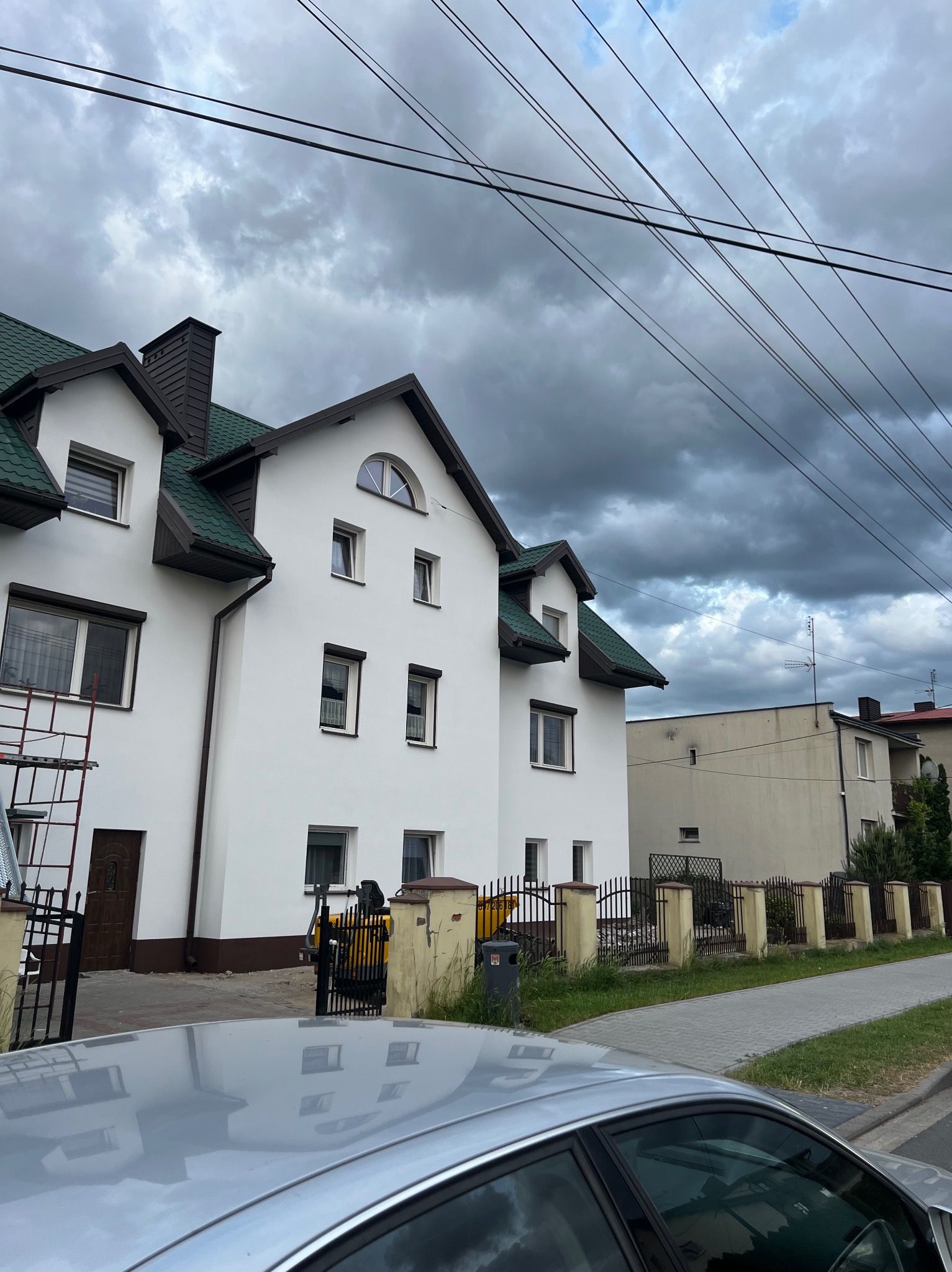 Biały dom z zielonym dachem po remoncie elewacji, widoczny zza samochodu, drabina przy ścianie, pochmurne niebo w tle.