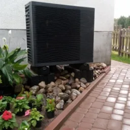 Czarna jednostka zewnętrzna urządzenia grzewczego Panasonic seria K, umieszczona na betonowych blokach przy ścianie domu, otoczona ozdobnymi kamieniami i różnorodną roślinnością w donicach. Obok...