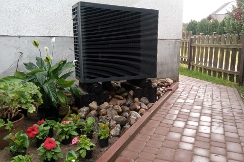 Czarna jednostka zewnętrzna urządzenia grzewczego Panasonic seria K, umieszczona na betonowych blokach przy ścianie domu, otoczona ozdobnymi kamieniami i różnorodną roślinnością w donicach. Obok...