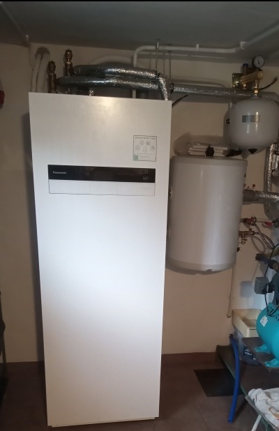 Biała jednostka wewnętrzna pompy ciepła Panasonic typu 'all in one', zintegrowana ze zbiornikiem na ciepłą wodę i elementami układu, widoczna na tle rur instalacji grzewczej i zbiornika...