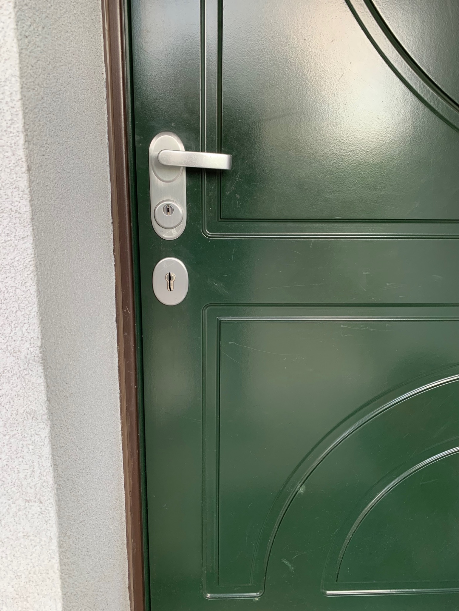 Zielone, metalowe drzwi wejściowe z srebrną klamką i zamkami, widoczne z bliska, z fragmentem ściany obok.