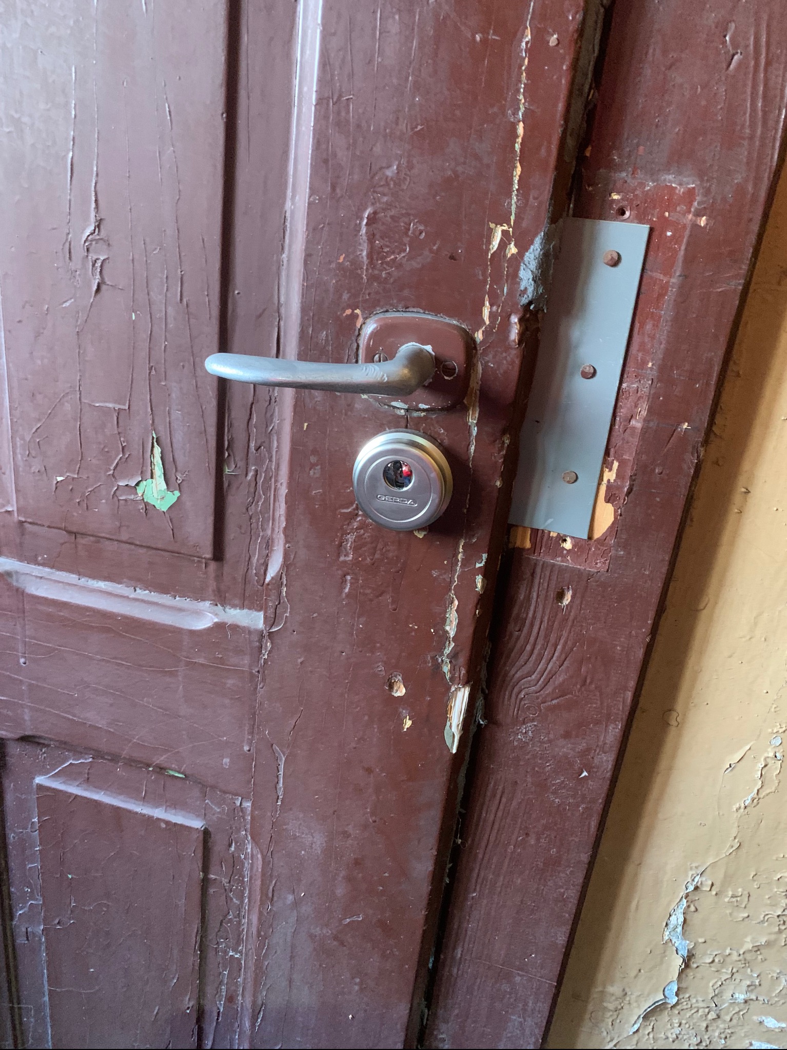 Stare, brązowe drzwi z zamontowanym nowym zamkiem i wzmocnieniem, widoczne odpryski farby i uszkodzenia drewna.