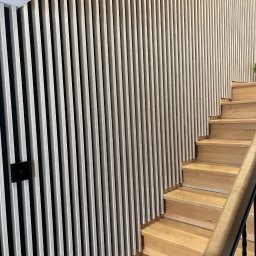 Tomasz Duda - Nowoczesne schody drewniane z balustradą i panelami ściennymi z pionowych listew w jasnym odcieniu drewna, z widocznym włącznikiem światła na ścianie.