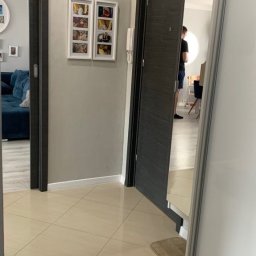 Tomasz Duda - Beżowe płytki podłogowe w przedpokoju z szarą ścianą, na której wiszą zdjęcia w białych ramkach, oraz ciemnoszare drzwi z napisem 'K+M+O 2021'.