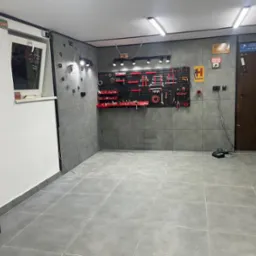 Wyposażony warsztat z szarymi płytkami na ścianach i podłodze, narzędzia na ścianie, zegar ścienny, szafka narzędziowa MEECTOOLS i oświetlenie sufitowe.