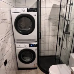 Kompaktowa łazienka z pralką i suszarką Electrolux ustawionymi jedna na drugiej, kabina prysznicowa z czarnymi profilami i biała toaleta.