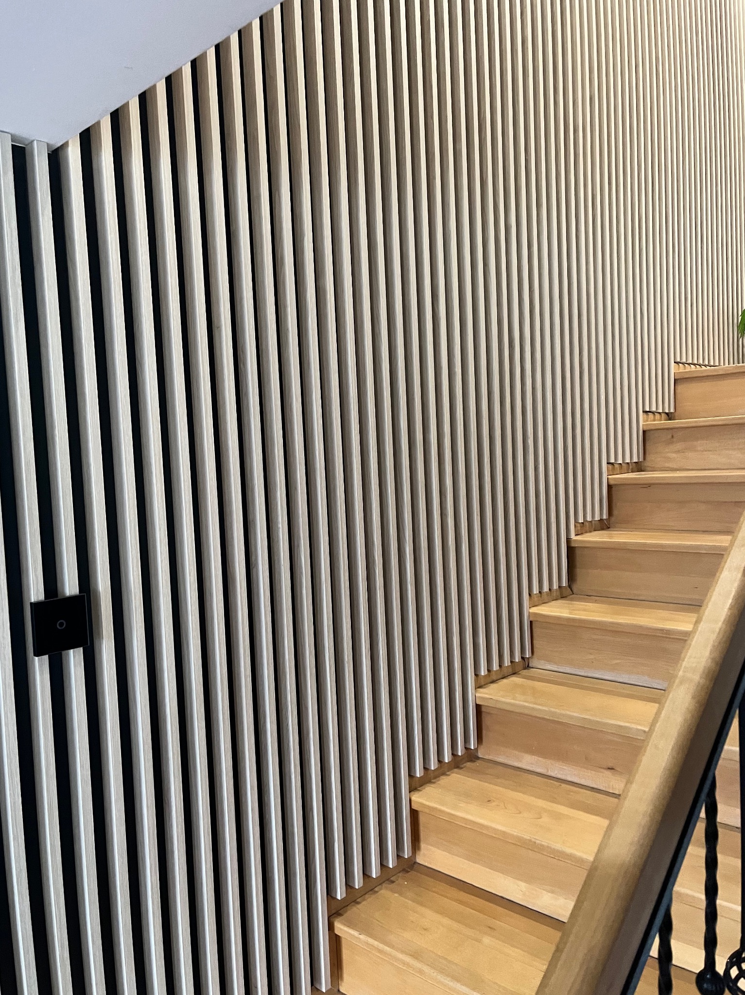 Nowoczesne schody drewniane z balustradą i panelami ściennymi z pionowych listew w jasnym odcieniu drewna, z widocznym włącznikiem światła na ścianie.