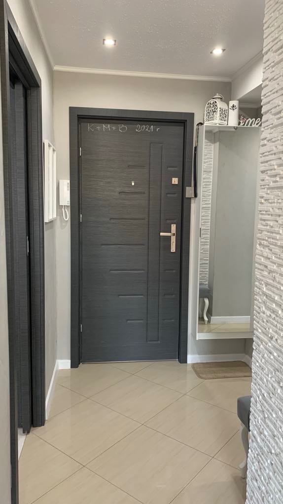 Nowoczesne drzwi wejściowe w kolorze grafitowym z poziomymi frezowaniami, zamontowane w jasnym przedpokoju z beżowymi płytkami podłogowymi i napisem 'K+M+B 2021 r' nad klamką.