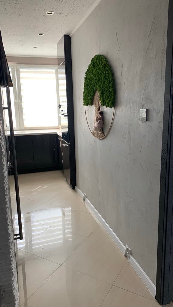 Korytarz z błyszczącą, beżową podłogą w kwadratowe płytki, szarą ścianą z dekoracją w kształcie drzewa z mchu i ciemnymi drzwiami prowadzącymi do kuchni z czarnymi szafkami.