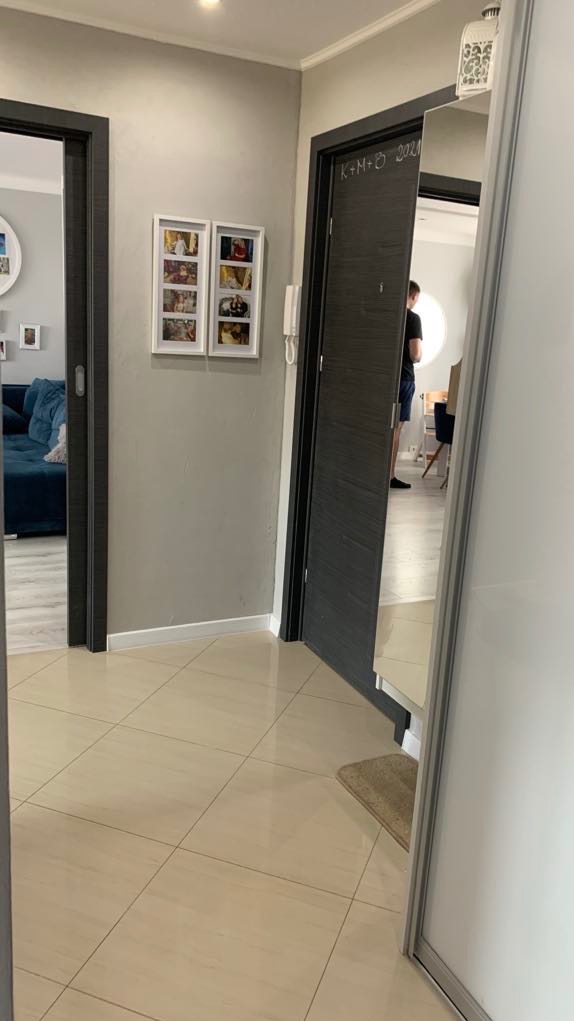 Beżowe płytki podłogowe w przedpokoju z szarą ścianą, na której wiszą zdjęcia w białych ramkach, oraz ciemnoszare drzwi z napisem 'K+M+O 2021'.