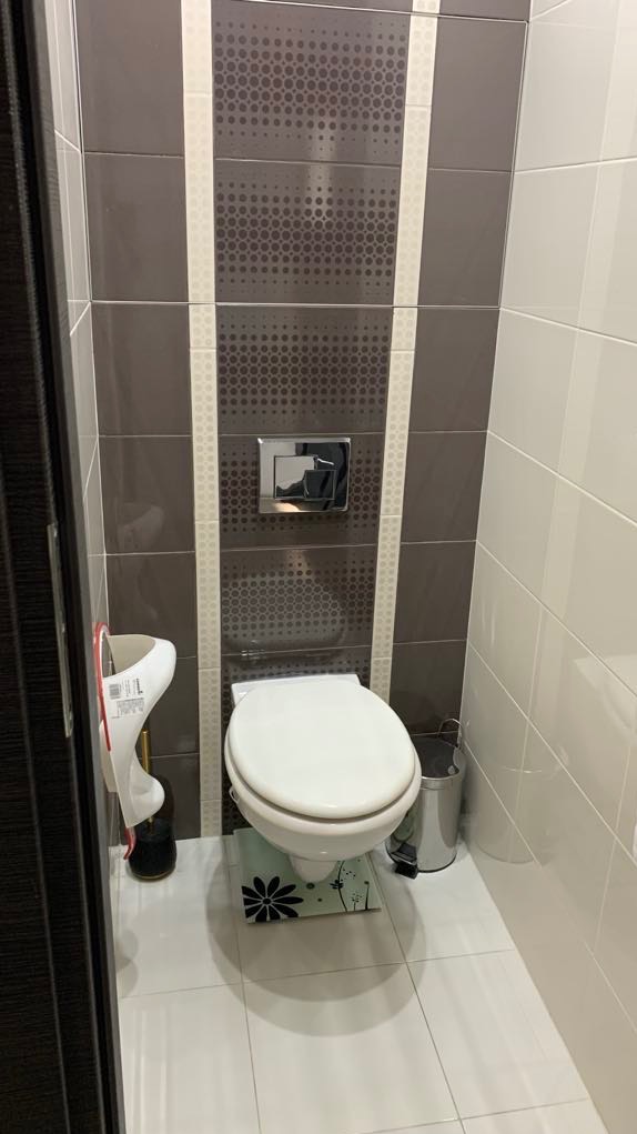 Wąska łazienka z brązowo-białymi płytkami z motywem kropek na ścianie za toaletą, białą toaletą i akcesoriami, widok z perspektywy drzwi.