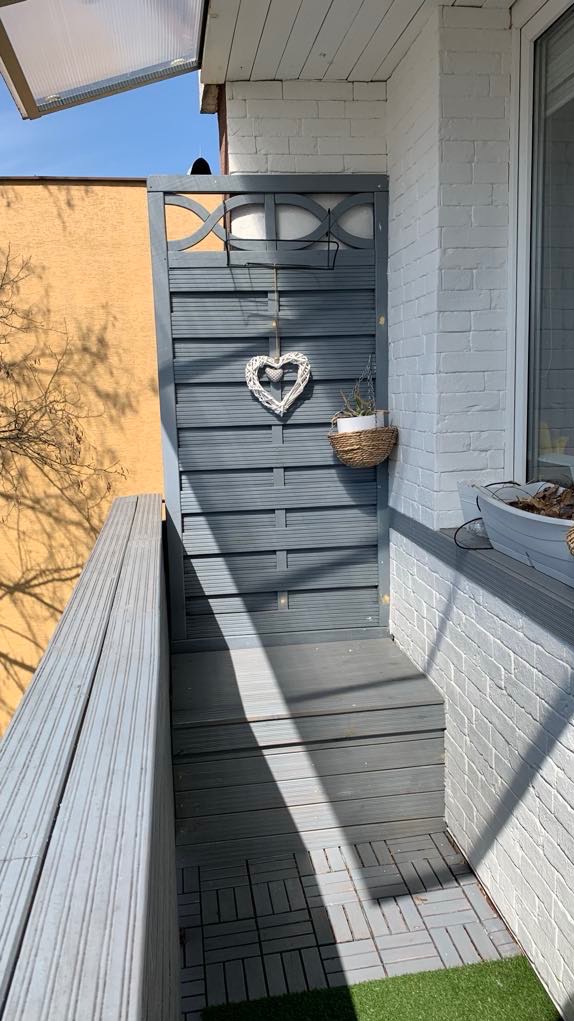 Szary balkon z drewnianą podłogą i ścianą z dekoracyjnym sercem i wiszącą doniczką, biała ceglana ściana z oknem po prawej stronie.