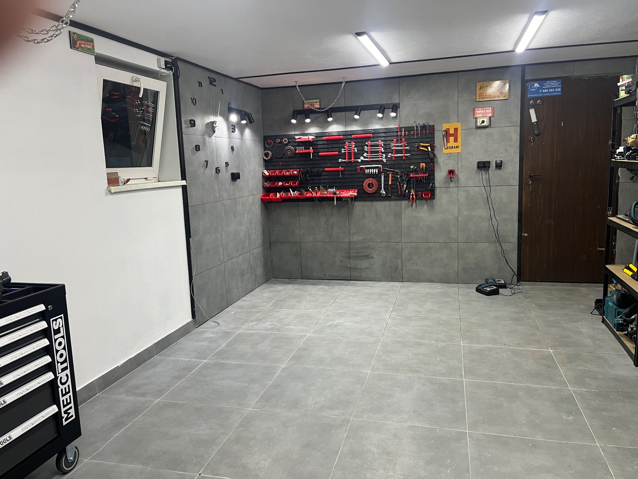 Wyposażony warsztat z szarymi płytkami na ścianach i podłodze, narzędzia na ścianie, zegar ścienny, szafka narzędziowa MEECTOOLS i oświetlenie sufitowe.