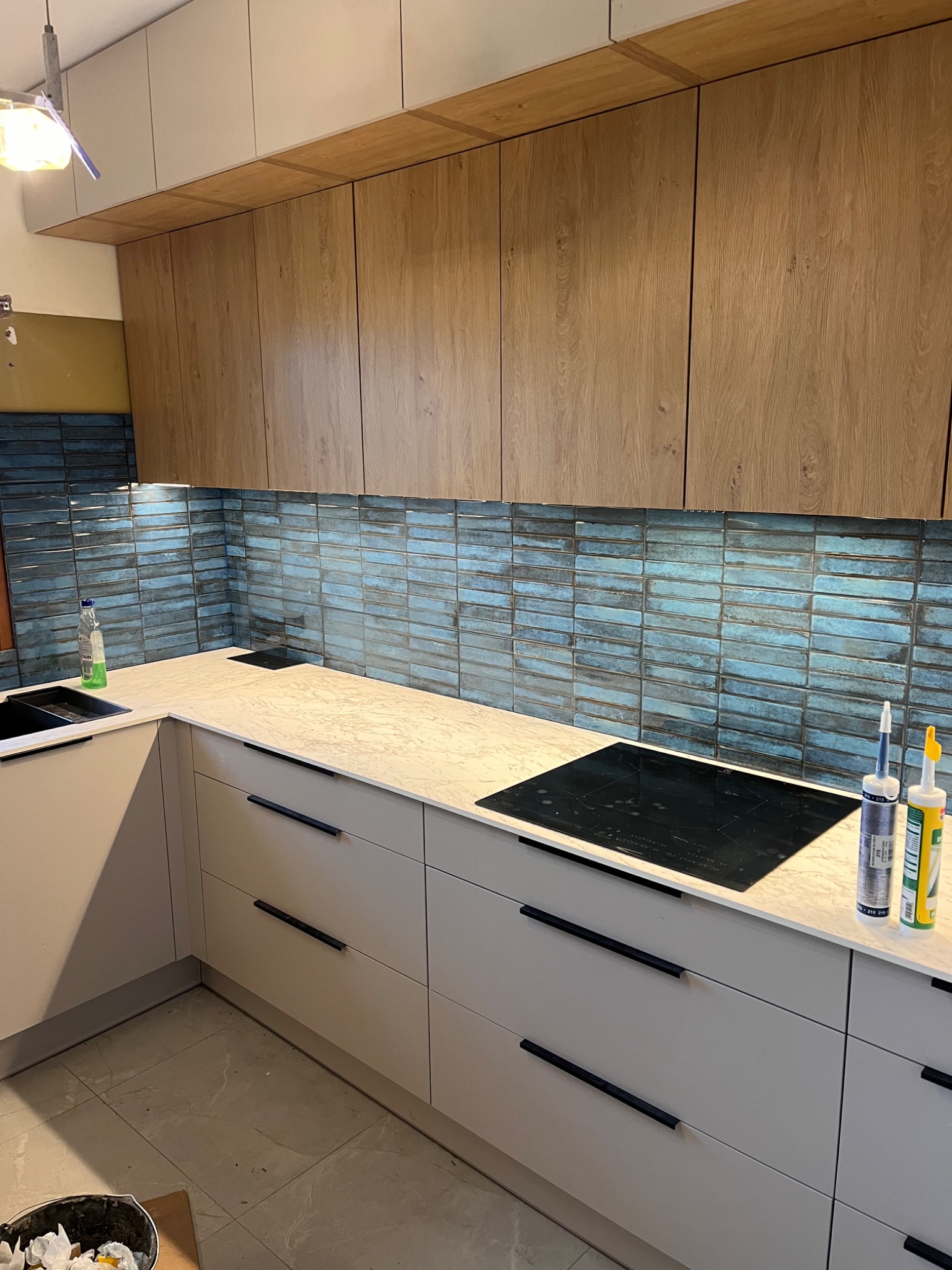 Nowoczesna kuchnia z drewnianymi szafkami wiszącymi, blat z efektem kamienia i niebieską, cegiełkową mozaiką na ścianie, uzupełniona czarną płytą indukcyjną i tubami z silikonem na blacie.