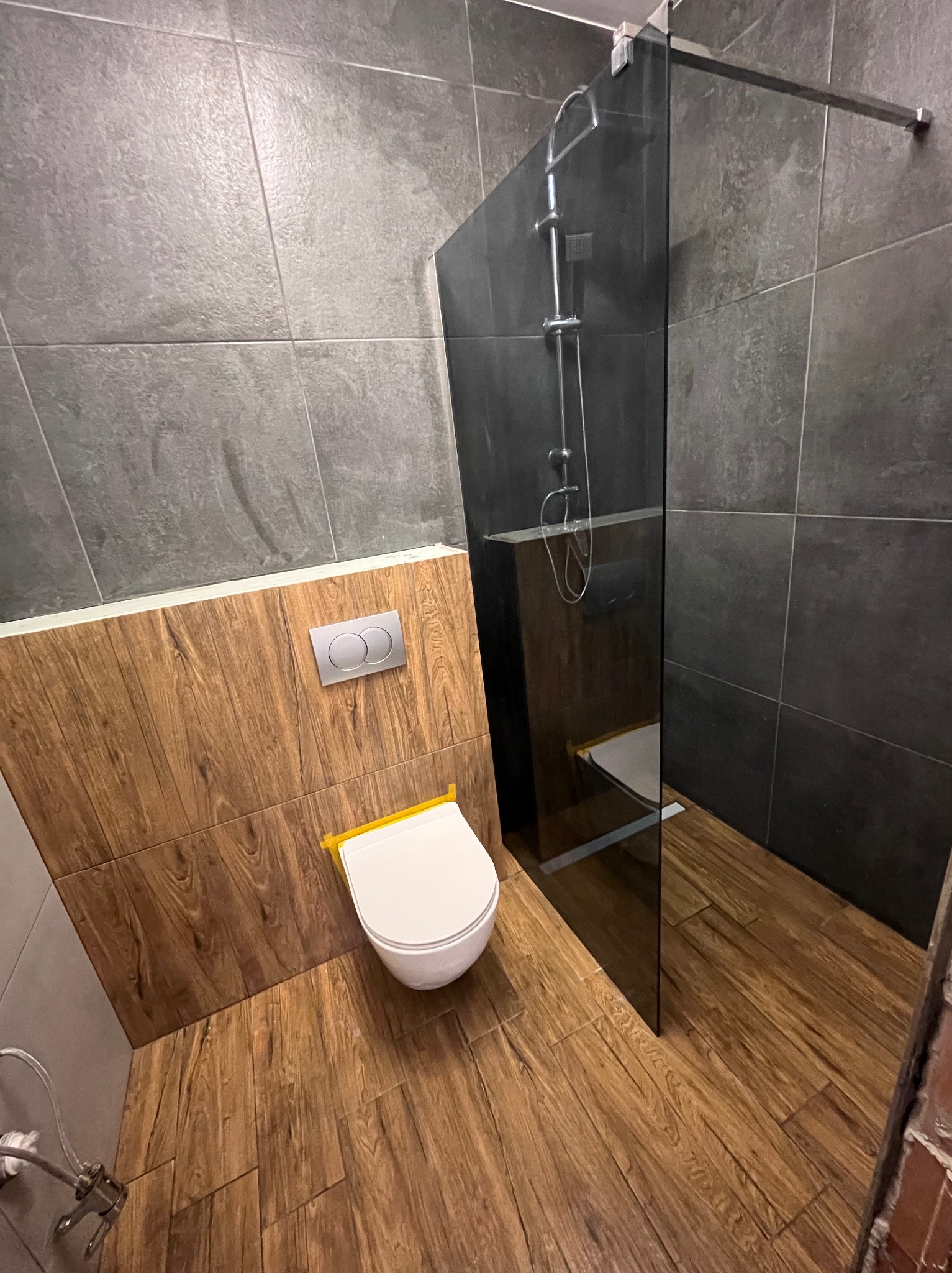 Nowoczesna łazienka z szarą płytką imitującą kamień na ścianach, drewnopodobną płytką na podłodze i ścianie za toaletą, szklaną kabiną prysznicową typu walk-in oraz wiszącą miską WC z chromowanym...