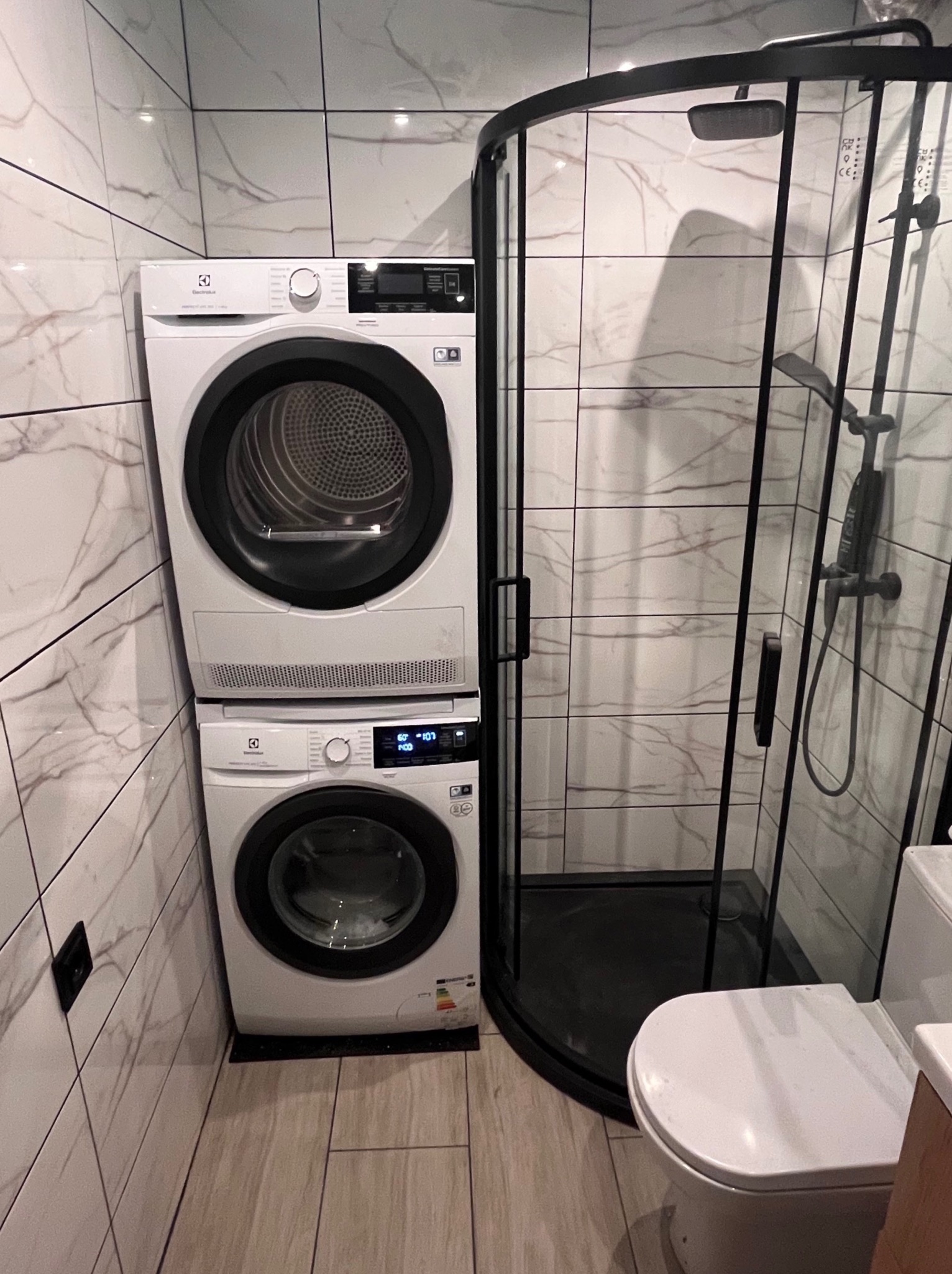Kompaktowa łazienka z pralką i suszarką Electrolux ustawionymi jedna na drugiej, kabina prysznicowa z czarnymi profilami i biała toaleta.