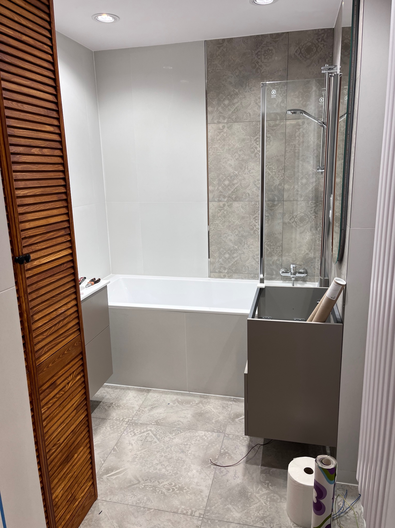 Nowoczesna łazienka po remoncie: szare płytki z wzorem, biała wanna, drewniane żaluzje, szklany parawan nawannowy i szara szafka pod umywalką. Widoczne akcesoria montażowe.