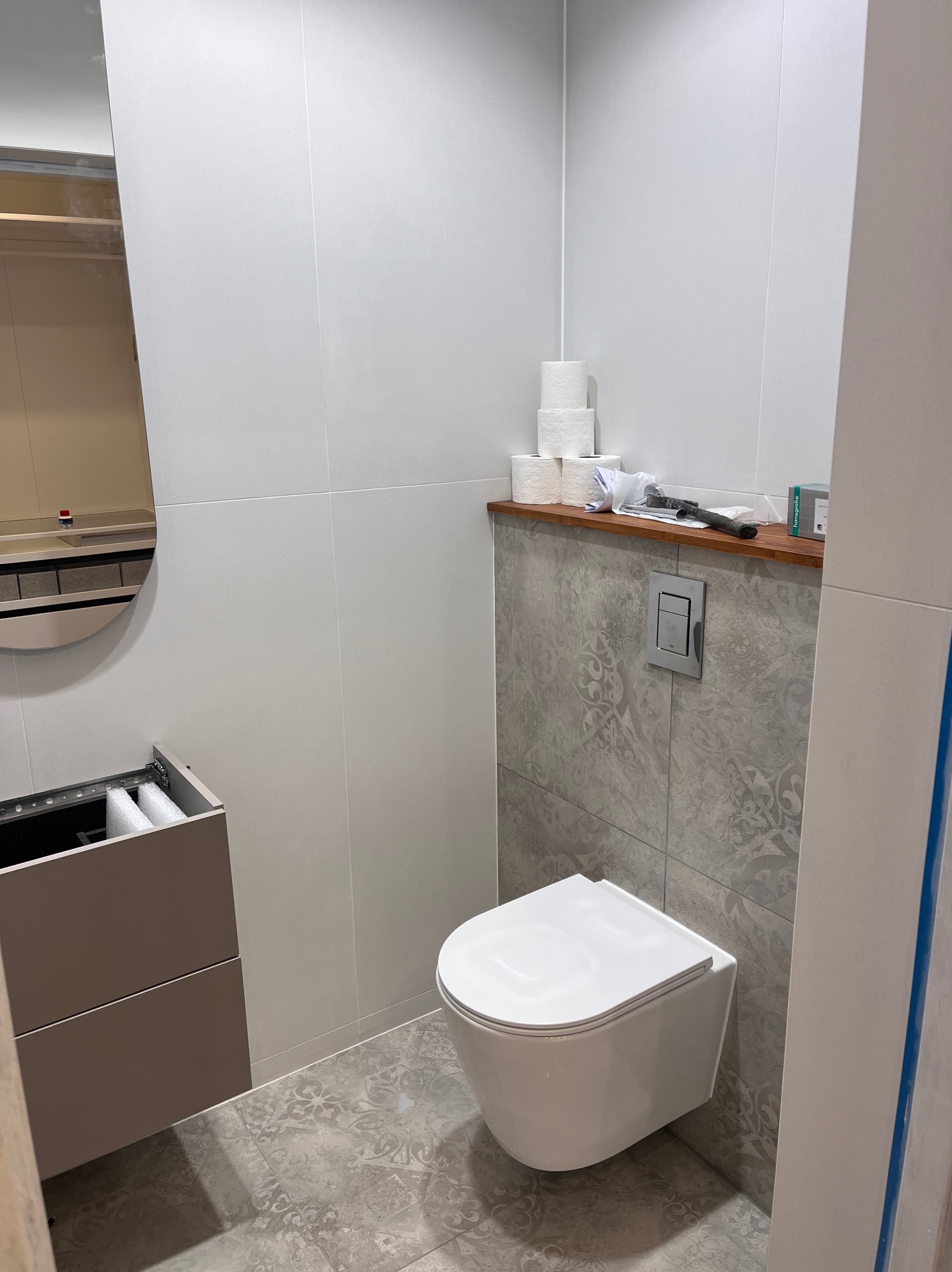 Nowoczesna łazienka w trakcie wykańczania: podwieszana toaleta, umywalka z lustrem, szare płytki z wzorem i białe ściany. Widoczne narzędzia i materiały.