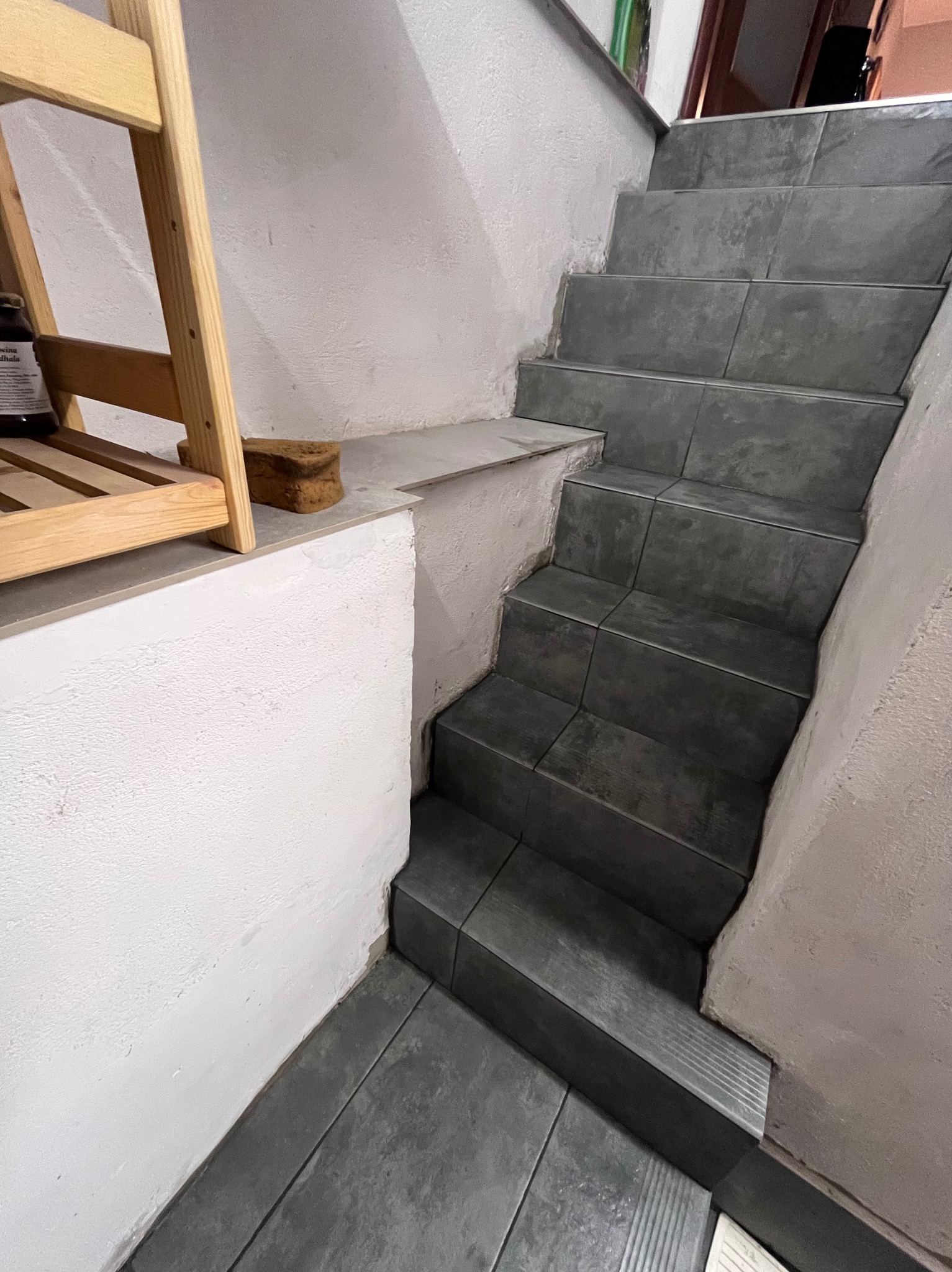 Wykończone płytkami schody w piwnicy, z półką i drewnianym regałem. Szare płytki na stopniach i podłodze, białe ściany. Ujęcie z dołu, podkreślające geometrię.