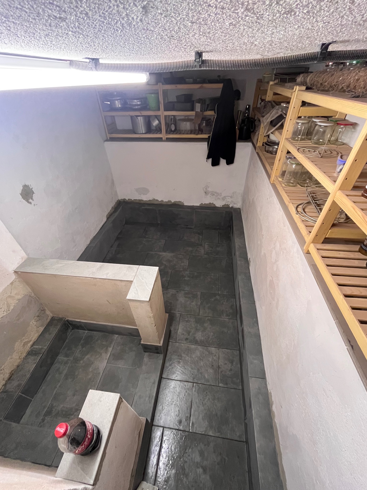 Ułożone ciemne płytki podłogowe w pomieszczeniu z surowymi ścianami. Widoczne półki z słoikami i butelka napoju. Prace wykończeniowe w piwnicy.