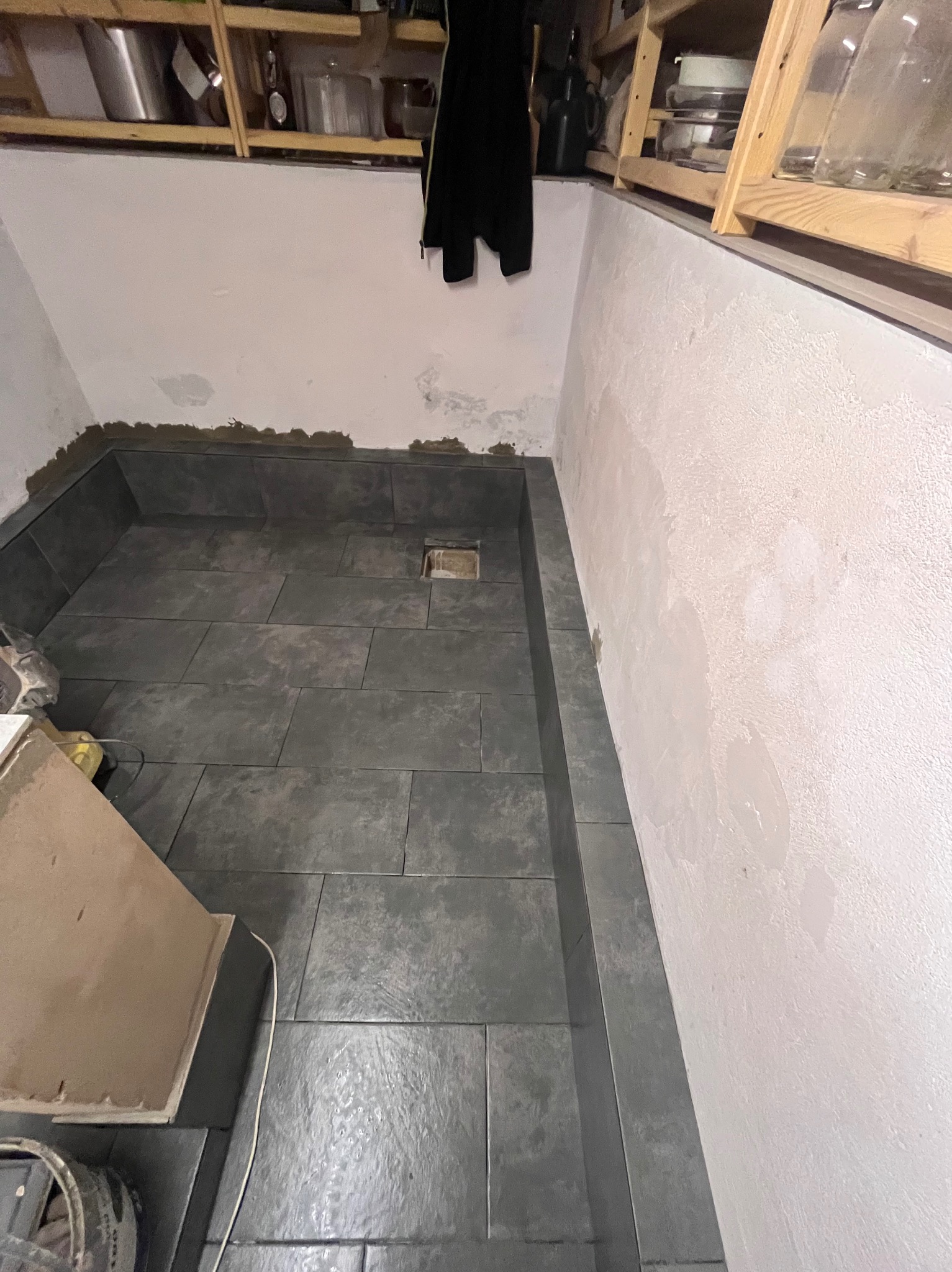 Ułożone, ciemne płytki podłogowe w łazience, widok z góry. Na ścianach widoczne ślady po remoncie. Półki z naczyniami w tle.