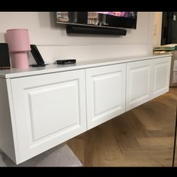 GPMeble Grzegorz Ptak - Biała, podwieszana szafka RTV z frezowanymi frontami, zamontowana pod telewizorem na ścianie. Parkiet ułożony w jodełkę. Element wystroju wnętrza.