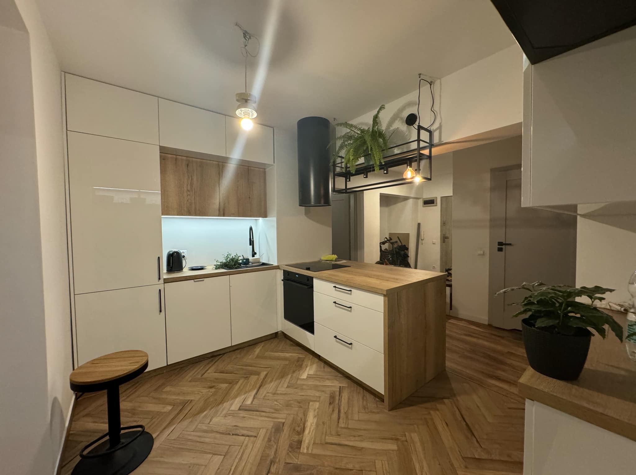 Nowoczesna kuchnia z wyspą i parkietem w jodełkę. Białe fronty szafek kontrastują z drewnianymi blatami. Nad wyspą designerski okap i półka na rośliny.