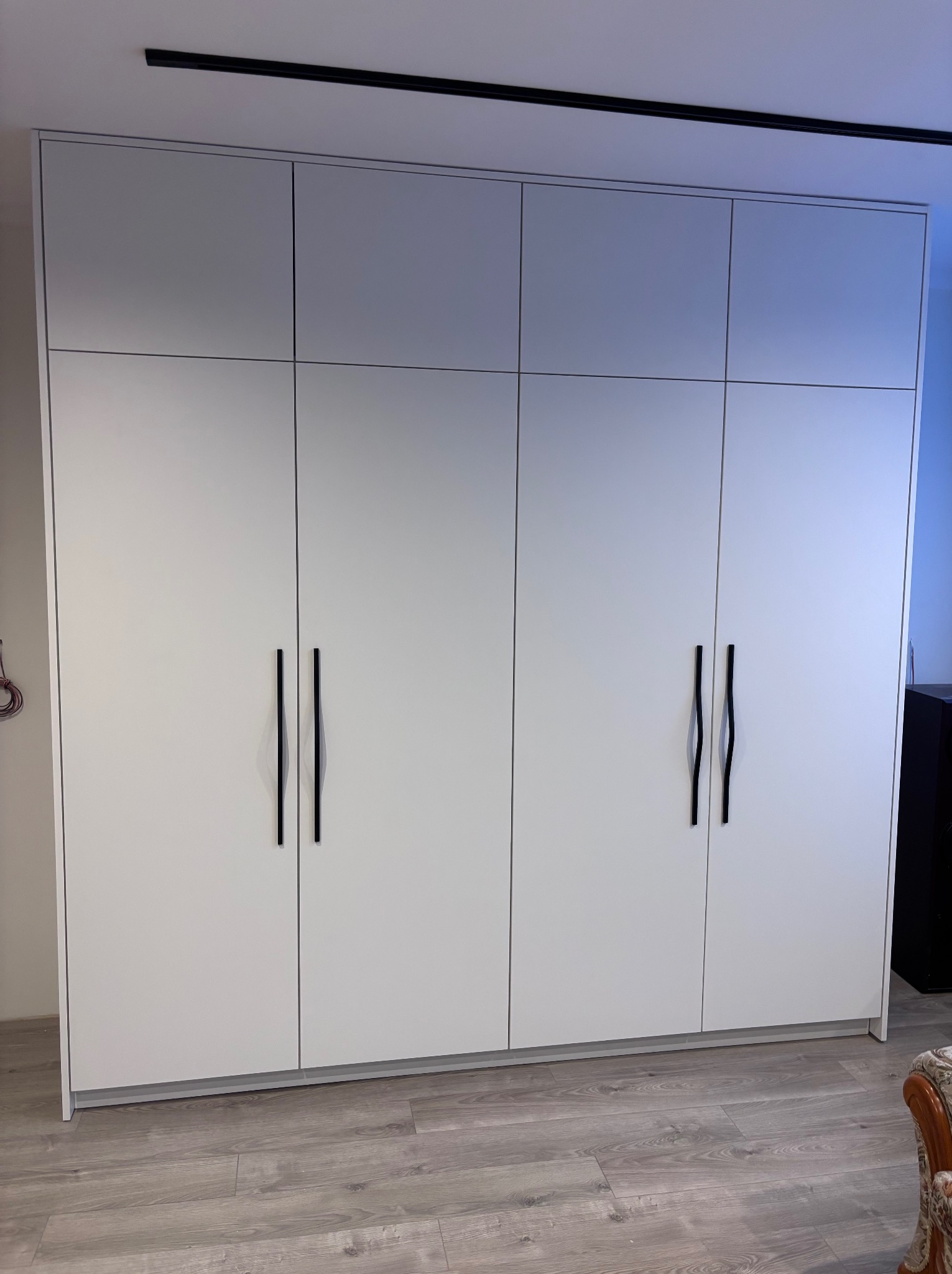 Biała, nowoczesna szafa wnękowa z czterema drzwiami i podłużnymi, czarnymi uchwytami, na tle paneli podłogowych i fragmentu ściany. Minimalistyczny design.