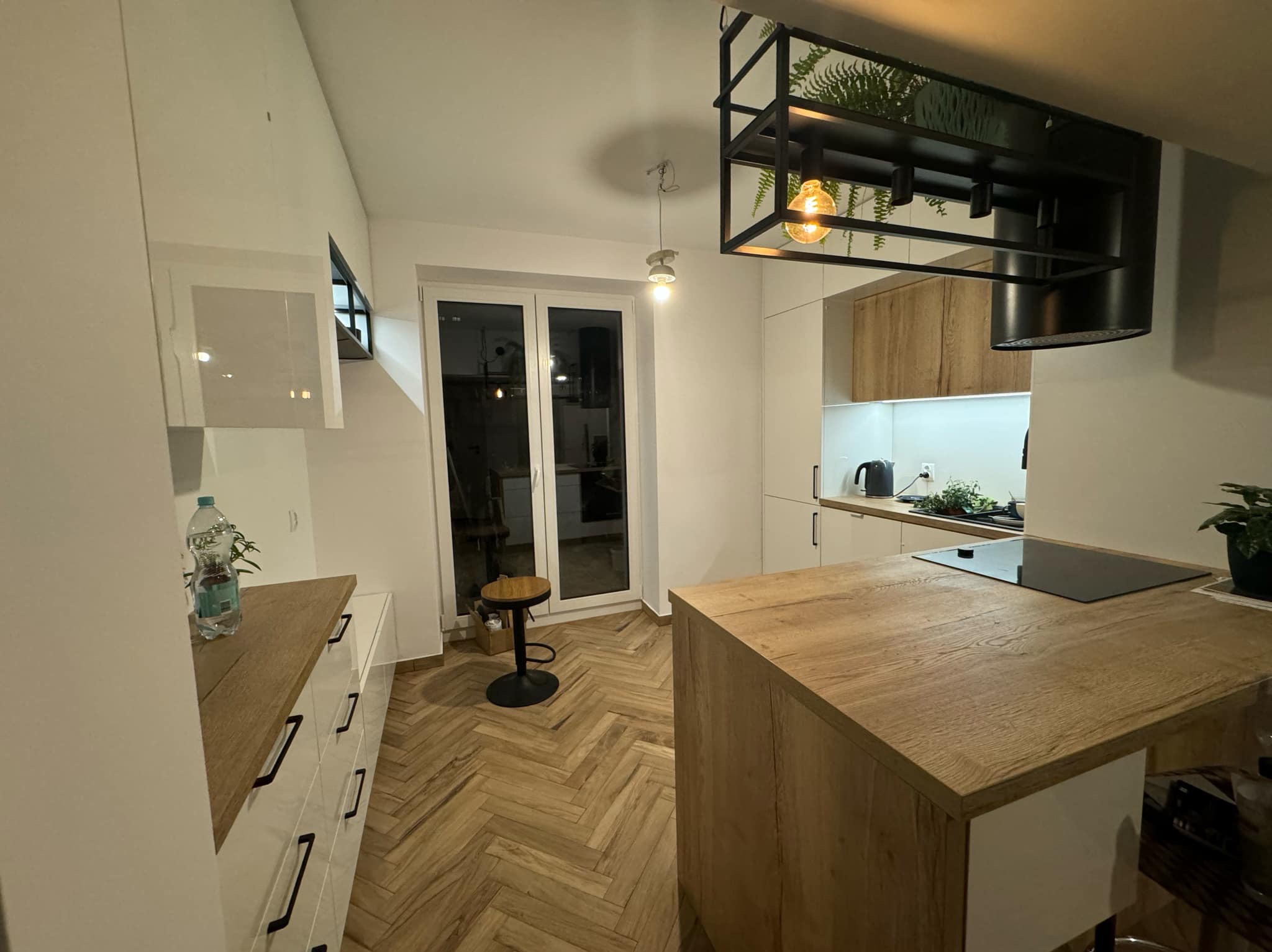 Jasna kuchnia z drewnianym blatem wyspy i parkietem w jodełkę. Nad wyspą designerska lampa z roślinami. Minimalistyczny styl z akcentami natury.